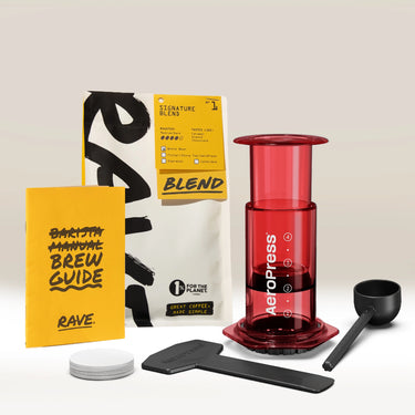 An Aeropress Gift Set Clear/Colour by Aeropress, perfect as a coffee gift or for any home brewer, includes a scoop, stirrer, filters, yellow Rave brew guide, and a bag of Rave Signature Blend coffee, all displayed on a light surface.
