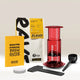 An Aeropress Gift Set Clear/Colour by Aeropress, perfect as a coffee gift or for any home brewer, includes a scoop, stirrer, filters, yellow Rave brew guide, and a bag of Rave Signature Blend coffee, all displayed on a light surface.