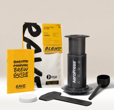 A coffee brewing kit arranged on a light beige studio background, including a black AeroPress, a yellow and white coffee bag, a yellow Rave manual brew guide, a black scoop, a black spatula, and small white lids.