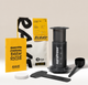 A coffee brewing kit arranged on a light beige studio background, including a black AeroPress, a yellow and white coffee bag, a yellow Rave manual brew guide, a black scoop, a black spatula, and small white lids.