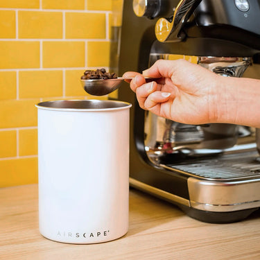 A hand uses the Planetary Design Airscape Coffee Scoop to collect beans from a stainless steel tin beside a black espresso machine on a kitchen counter with a yellow tile backsplash.