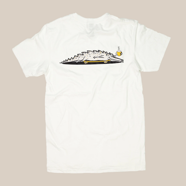 The Alligator | T-Shirt by RAVE COFFEE is a white unisex cotton tee featuring a cartoon crocodile skateboarding with coffee in hand and a relaxed vibe, set against a plain beige background.
