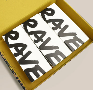 Three white rectangular boxes with large black brushstroke-style letters spelling 'RAVE' are arranged inside a mustard yellow shipping box on a light beige background.