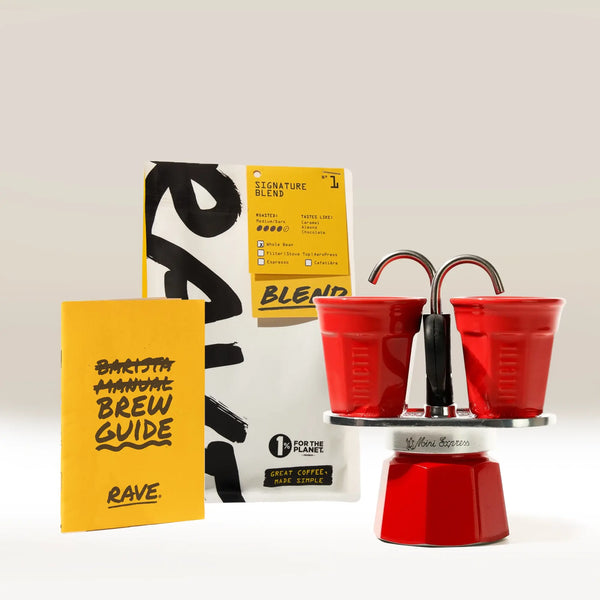 A Bialetti Mini Express Stove Top Gift Set, featuring a red coffee maker with two matching cups, is displayed alongside a yellow brew guide booklet and a white-and-yellow bag of Rave Signature Blend espresso coffee on a light background.