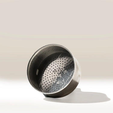 A Bialetti Funnel for Ground Coffee by Bialetti, resembling a metal espresso machine filter basket, lies on its side on a light surface with its fine mesh interior and engraved text visible, casting a shadow.