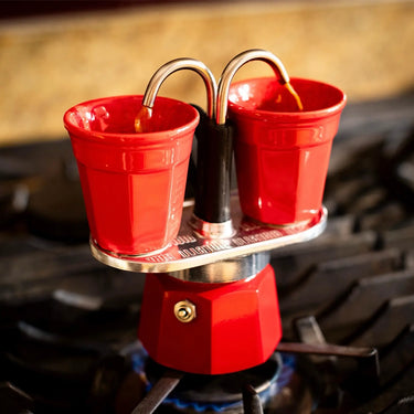 The Bialetti Mini Express Stove Top Gift Set by Bialetti brews espresso directly on your stove, elegantly pouring rich coffee through two spouts into a pair of matching red cups placed on top.