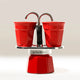 A Bialetti Mini Express Moka Pot in red, with two matching red cups on top to catch coffee from the curved spouts. The background is plain and light-colored.
