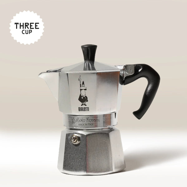 A Bialetti Moka Express Stove Top Coffee Maker in silver, featuring a black handle, is displayed against a plain background. A "THREE CUP" label appears in the top left corner.
