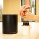 A person uses an AeroPress over a white mug beside the Planetary Design Black Airscape Coffee Storage Container on a light wood table, with windows and trees in the background.