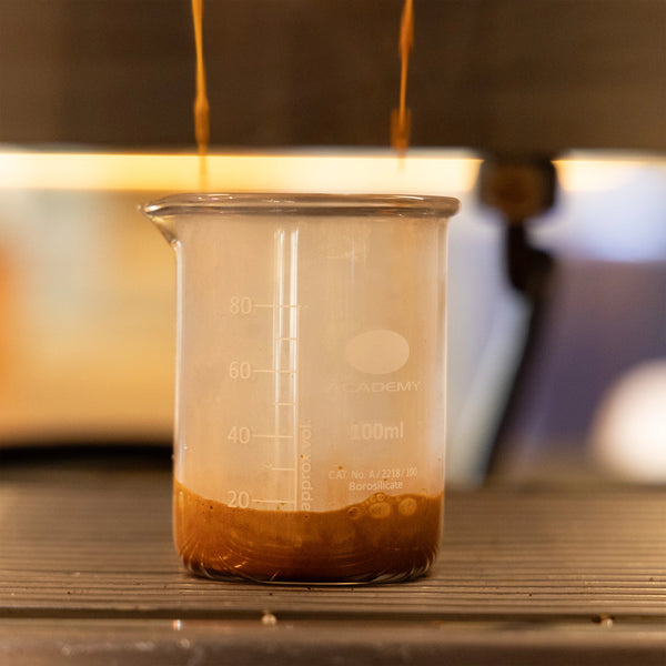 A close-up of espresso pouring into a 100ml Labtek Borosilicate Glass Beaker on a coffee machine tray—the perfect shot catcher for precise, mess-free pours.