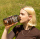 A person with short blonde hair drinks from a large brown RAVE COFFEE Rave Nalgene Bottle (1L) labeled Not Coffee while standing outdoors on a sunny day with green grass in the background.