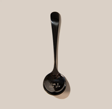 A shiny black stainless steel Rave Cupping Spoon from RAVE Coffee rests on a beige background, with “CAN I SIP IT?” engraved on the bowl—a playful nod to the art of coffee tasting and cupping.