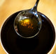 A RAVE Coffee Rave Cupping Spoon hovers above a cup of dark liquid, its stainless steel surface reflecting the words CAN I SIP IT?—a playful tribute to the art of coffee tasting and cupping.