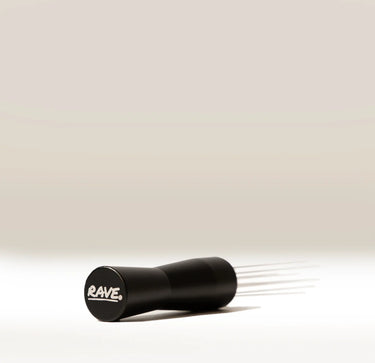 A black RAVE brand WDT tool with multiple thin metal prongs extending from one end, shown on a clean, light beige-to-white gradient background.