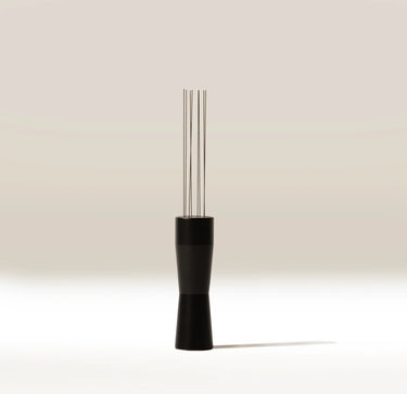 A modern coffee tool with a matte black handle and four slender stainless steel wires, standing upright on a clean white surface against a warm beige background.