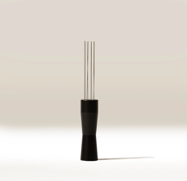 A modern coffee tool with a matte black handle and four slender stainless steel wires, standing upright on a clean white surface against a warm beige background.