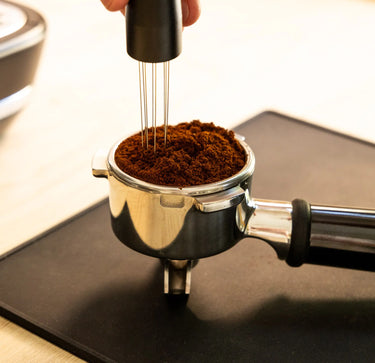 A hand uses the RAVE Coffee WDT Tool to stir ground coffee in a portafilter, ensuring even distribution of the grounds and preparing them for brewing espresso on a black mat.