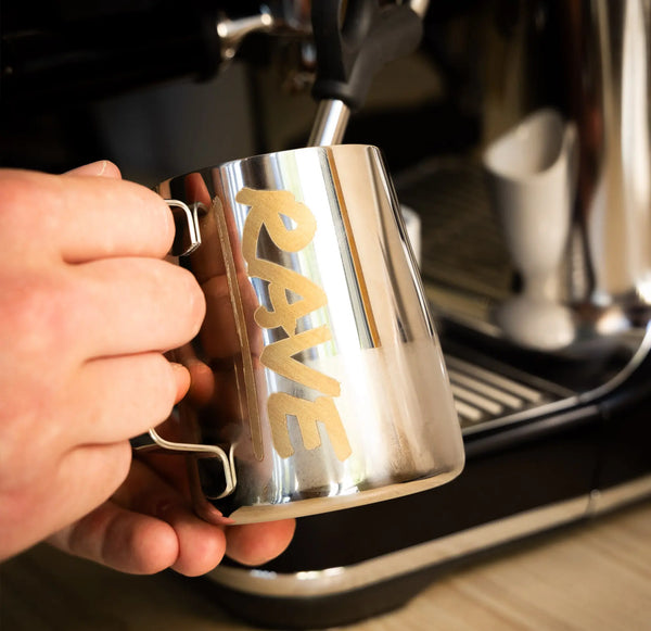 A hand holds the Rave Milk Jug from RAVE COFFEE under a steam wand of an espresso machine, preparing to steam milk into perfect microfoam.