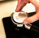 A close-up of a hand holding a metal coffee portafilter with the words '50 FRESH 50 CLEAN' engraved on the lid, positioned over a black tamper on a light wood surface.