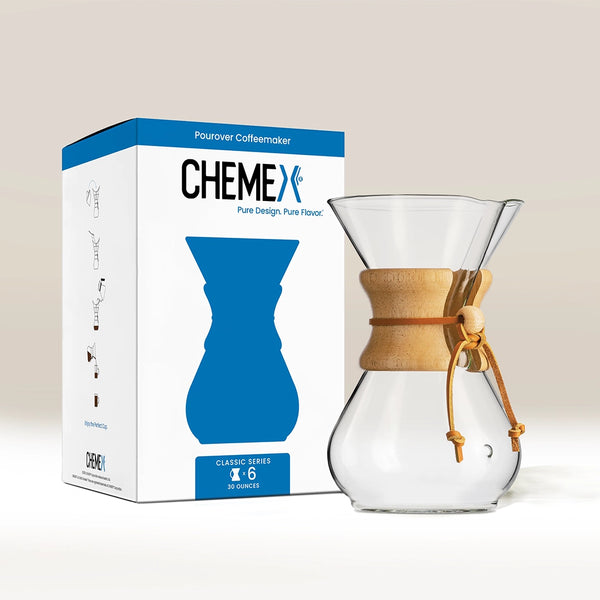 The Chemex (6 Cup) Classic, made from durable borosilicate glass with a wooden collar and leather tie, sits beside its white and blue box featuring the Chemex brand name and an illustration of this stylish home coffee maker.