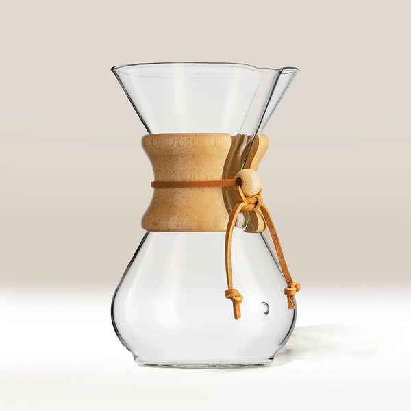 The Chemex (6 Cup) Classic by Chemex features clear borosilicate glass, a wooden collar, and a leather tie. Shown against a neutral background, it makes an elegant addition to any home coffee setup.