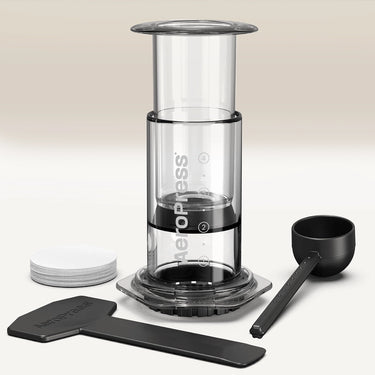 A Clear/Coloured AeroPress Coffee Maker by Aeropress stands upright on a light surface, with round paper filters, a black scoop, stirrer, and filter holder—everything included for portable coffee brewing—set against a neutral background.