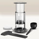 A Clear/Coloured AeroPress Coffee Maker by Aeropress stands upright on a light surface, with round paper filters, a black scoop, stirrer, and filter holder—everything included for portable coffee brewing—set against a neutral background.