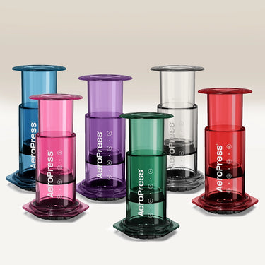 Six Aeropress Clear / Coloured Coffee Makers in blue, pink, purple, green, clear, and red are displayed in two rows on a neutral background. Each portable maker features the Aeropress logo on the side.