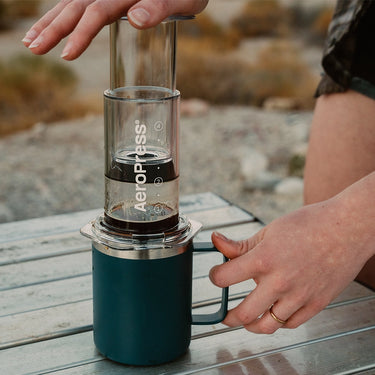 Outdoors, a person brews coffee using the Aeropress Clear / Coloured AeroPress Coffee Maker, pressing the plunger with one hand while holding a blue mug steady on a wooden surface.