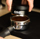 A hand uses the RAVE Coffee Rave 54mm Espresso Distributor to evenly distribute and level ground coffee in a 54mm portafilter, preparing it for optimal espresso extraction.