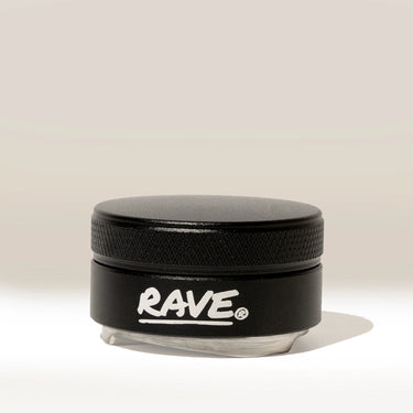 A small black RAVE Coffee container with a textured lid and “RAVE” written in white on the front, identified as the Rave 54mm Espresso Distributor and resembling an espresso distribution tool or coffee bed leveler, is placed against a plain, light background.