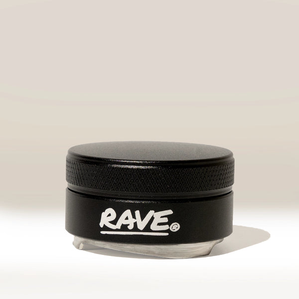 A small black RAVE Coffee container with a textured lid and “RAVE” written in white on the front, identified as the Rave 54mm Espresso Distributor and resembling an espresso distribution tool or coffee bed leveler, is placed against a plain, light background.