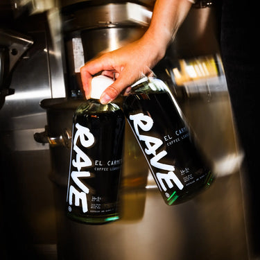 Barista holding a bottle of Rave El Carmen Coffee Liqueur beside an espresso machine.