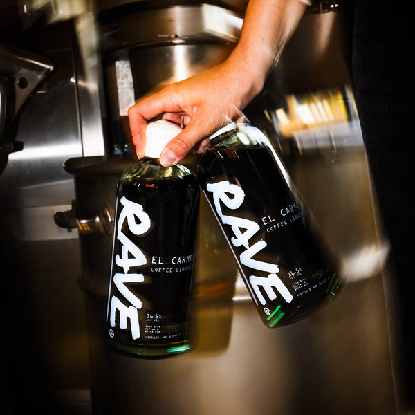 Barista holding a bottle of Rave El Carmen Coffee Liqueur beside an espresso machine.