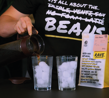 A photo of a person pouring an iced coffee from a glass coffee brewing mug into a two glasses with ice.