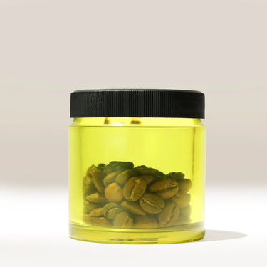 A well-lit, neutral background showcases the Comandante Polymer Bean Jar - Yellow by Comandante. The transparent jar with a black lid is half-filled with yellow liquid and whole coffee beans, ideal as a Comandante C40 accessory.