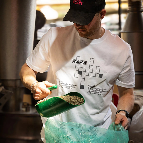 Person pouring coffee beans from a green scoop into a large green container, wearing a white t-shirt with text and graphics.