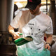 Person pouring coffee beans from a green scoop into a large green container, wearing a white t-shirt with text and graphics.
