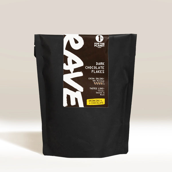 A black bag labeled RAVE COFFEE Drinking Chocolate Flakes stands upright. Bold white text and a yellow section highlight this hot chocolate. A 1% For The Planet logo sits at the top—ideal for fans of dark, milk, or white chocolate.