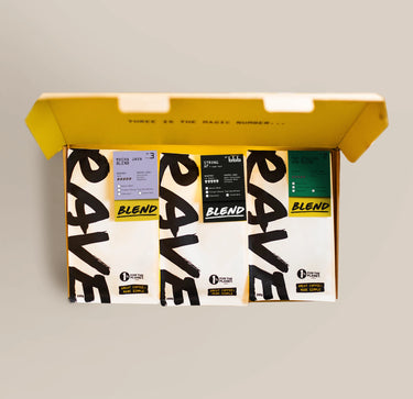 A Ravecoffee Dark Roast Bundle, featuring an open box containing three coffee bags with uniquely colored labels—purple, black, and green—each showcasing distinct blend details such as strong coffee and dark roast options, set against a plain background.