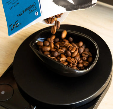 Roasted coffee beans pouring from a blue and white bag into a glossy black bowl placed on a digital scale.