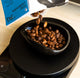 Roasted coffee beans pouring from a blue and white bag into a glossy black bowl placed on a digital scale.