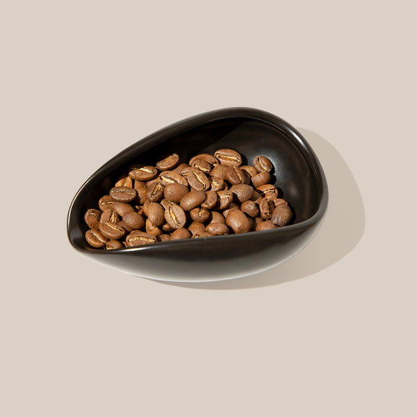 Roasted coffee beans in a glossy black teardrop-shaped bowl, set against a solid beige background.