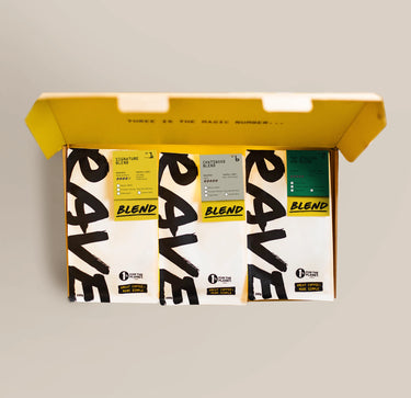 An open box reveals four bags of Ravecoffee’s Espresso Lover Coffee Bundle, each sporting a distinctively colored label—ideal for exploring and finding your new favorite espresso blend. Inside the lid, “Three is the magic number…” is printed in small text, suggesting exceptional flavors for both milk-based espresso drinks and bold dark roasts. The box sits against a plain beige background.