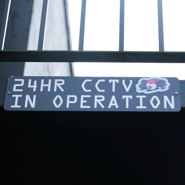 A black sign with white text reads 24HR CCTV IN OPERATION and features a small Rave Tattoo Sticker Pack holographic cartoon clown face in the top right corner. Metal bars and part of a building are visible above.