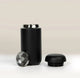 A matte black Fellow Carter Move Mug (12oz) with its lid removed and a metal splash guard standing next to it.