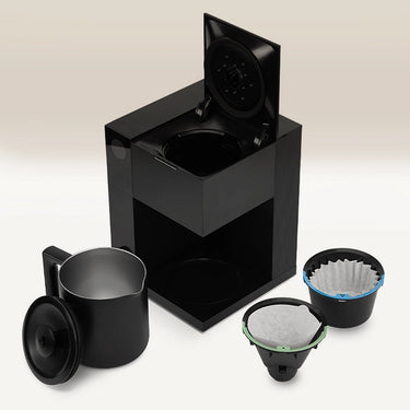 The Fellow Aiden Precision Drip Filter Coffee Maker by Fellow, shown in black with its lid open beside a matching carafe and two filter baskets, offers customizable temperature controls for the perfect brew, all arranged on a light background.