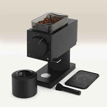 The Fellow Ode Brew Grinder Gen 2 by Fellow, with clear hopper holding coffee beans, sits beside a black knock box, a cleaning brush, and a black tray with measurement markings on a white surface.