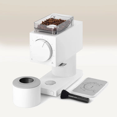 The Fellow Ode Brew Grinder Gen 2 by Fellow, shown in modern white with a clear bean-filled hopper, a round grounds container, a small cleaning brush, and a metal tray with measurement markings, is arranged on a neutral background.