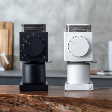 Two Fellow Ode Brew Grinder Gen 2 units—one black, one white—sit side by side on a wooden counter, each featuring Gen 2 Brew Burrs, a large front dial, and minimalist design with a blurred kitchen background.